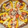 A keto pesto pepperoni pizza with a golden-brown crust. The pizza is topped with vibrant green pesto sauce, melted mozzarella cheese, circular slices of pepperoni, chopped mushrooms, and diced tomatoes. Curved strips of yellow bell pepper add a visually appealing contrast of colors. The pizza is cut into eight equal slices and placed on parchment paper on a baking tray, suggesting it is freshly baked and ready to serve.