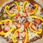 A keto pesto pepperoni pizza with a golden-brown crust. The pizza is topped with vibrant green pesto sauce, melted mozzarella cheese, circular slices of pepperoni, chopped mushrooms, and diced tomatoes. Curved strips of yellow bell pepper add a visually appealing contrast of colors. The pizza is cut into eight equal slices and placed on parchment paper on a baking tray, suggesting it is freshly baked and ready to serve.