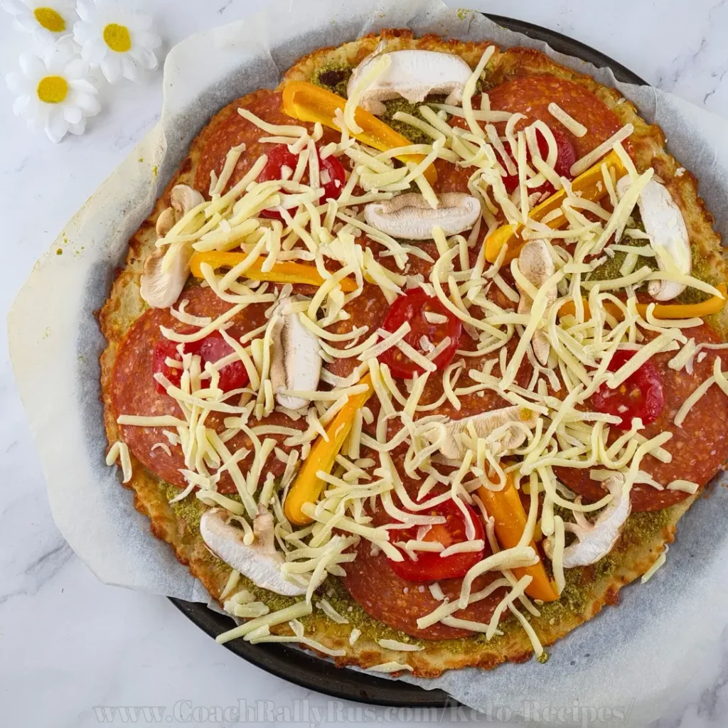 A keto pesto pepperoni pizza with a golden-brown crust. The pizza is topped with vibrant green pesto sauce, melted mozzarella cheese, circular slices of pepperoni, chopped mushrooms, and diced tomatoes. Curved strips of yellow bell pepper add a visually appealing contrast of colors. The pizza is cut into eight equal slices and placed on parchment paper on a baking tray, suggesting it is freshly baked and ready to serve.