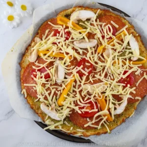 A keto pesto pepperoni pizza with a golden-brown crust. The pizza is topped with vibrant green pesto sauce, melted mozzarella cheese, circular slices of pepperoni, chopped mushrooms, and diced tomatoes. Curved strips of yellow bell pepper add a visually appealing contrast of colors. The pizza is cut into eight equal slices and placed on parchment paper on a baking tray, suggesting it is freshly baked and ready to serve.
