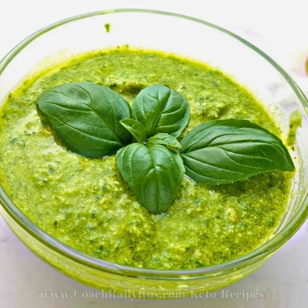 A vibrant green keto pesto sauce in a clear glass bowl, garnished with fresh basil leaves on top. The pesto’s texture appears creamy with visible flecks of herbs and nuts, indicating its fresh ingredients. The background is softly lit, highlighting the freshness and homemade quality of the sauce.