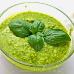 A vibrant green keto pesto sauce in a clear glass bowl, garnished with fresh basil leaves on top. The pesto’s texture appears creamy with visible flecks of herbs and nuts, indicating its fresh ingredients. The background is softly lit, highlighting the freshness and homemade quality of the sauce.