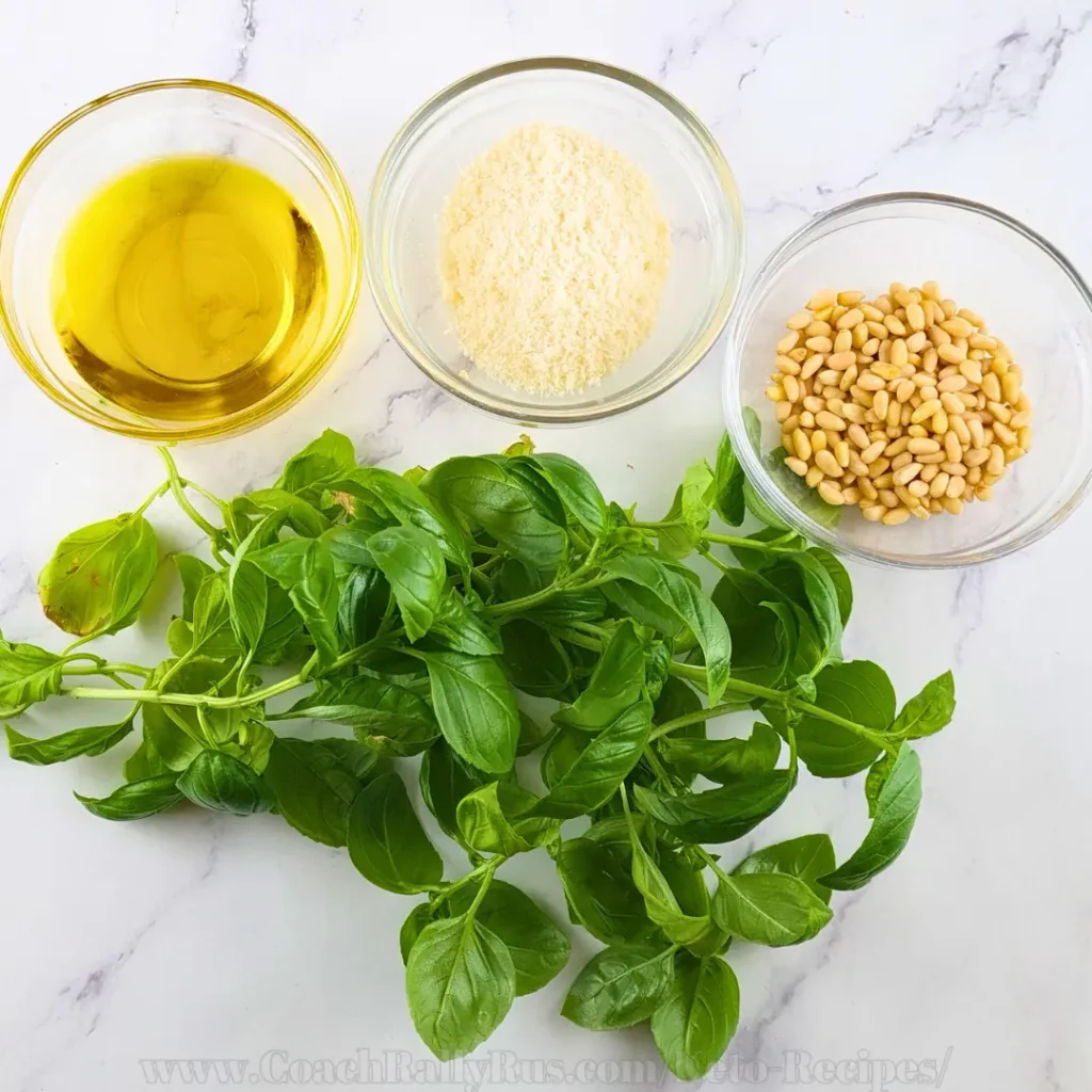 An overhead image of ingredients for keto pesto sauce arranged on a marble surface. Clockwise from the top left, there is a small bowl of olive oil, a bowl of grated Parmesan cheese, and a bowl of pine nuts. At the bottom center, there is a large bunch of fresh basil leaves. The ingredients are neatly portioned and presented, suggesting preparation for making pesto sauce.