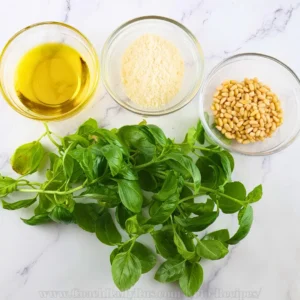 An overhead image of ingredients for keto pesto sauce arranged on a marble surface. Clockwise from the top left, there is a small bowl of olive oil, a bowl of grated Parmesan cheese, and a bowl of pine nuts. At the bottom center, there is a large bunch of fresh basil leaves. The ingredients are neatly portioned and presented, suggesting preparation for making pesto sauce.