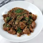 A plate of keto chicken liver, garnished with herbs, accompanied by a fork and knife, and a bowl of sauce on a marble surface.
