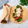 A photo of a keto chicken wrap with ranch dressing on a marble countertop. The wrap is made of a light brown tortilla filled with chicken, spinach, and a white sauce. The chicken is cooked and seasoned, the spinach is fresh and green, and the white sauce is creamy and tangy. There is a bunch of parsley and a gray and white striped towel in the background.