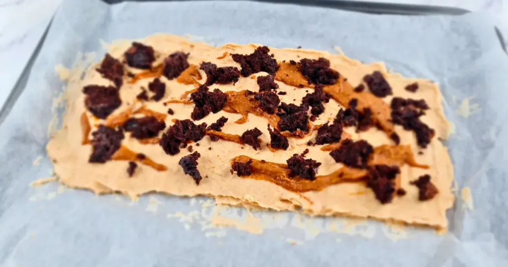This is a photo of a frozen yoghurt bark with peanut butter and brownie. The bark is rectangular in shape and has a light brown color. It is topped with crumbled brownie and chocolate chips. The bark is cut into smaller pieces and is on a white parchment paper on a white marble countertop.