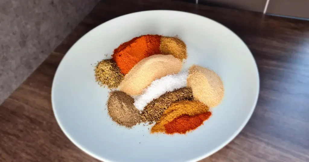 Keto Cajun Seasoning ingredients, including salt, garlic powder, onion powder, paprika, oregano, thyme, black pepper, cumin, coriander, and cayenne pepper, in a white bowl on a dark table.