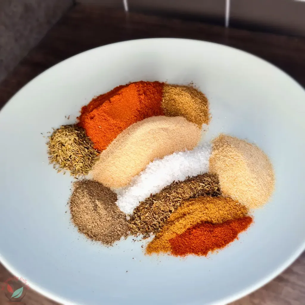 Keto Cajun Seasoning ingredients, including salt, garlic powder, onion powder, paprika, oregano, thyme, black pepper, cumin, coriander, and cayenne pepper, in a white bowl on a dark table.