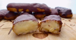 A close-up image of Keto Chocolate Eclairs displayed on a wooden surface. One eclair is cut in half, exposing the creamy filling inside, while others remain whole with a glossy chocolate coating.