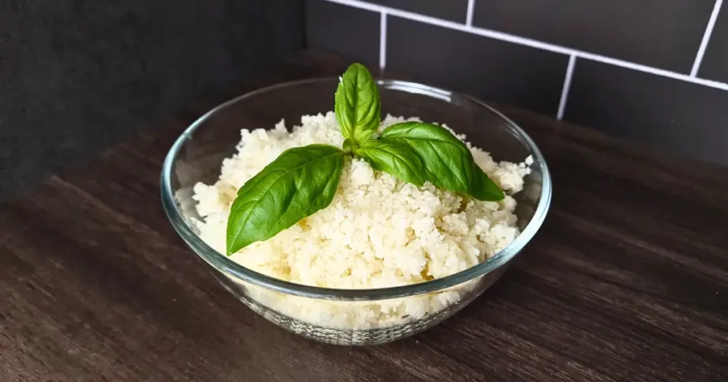 A photo of a glass bowl filled with green cauliflower rice, garnished with fresh basil leaves. The bowl is on a wooden countertop with a gray tile backsplash. The broccoli rice is a low-carb and keto-friendly alternative to regular rice, made by grating or processing cauliflower into rice-like grains.