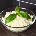 A photo of a glass bowl filled with green cauliflower rice, garnished with fresh basil leaves. The bowl is on a wooden countertop with a gray tile backsplash. The broccoli rice is a low-carb and keto-friendly alternative to regular rice, made by grating or processing cauliflower into rice-like grains.