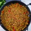 A photo of a black pan filled with a colorful and appetizing dish of Keto Mediterranean Cauliflower & Broccoli Rice, sprinkled with red pepper and onions. The dish is made with cauliflower and broccoli rice, onion, garlic, olive oil, basil, thyme, salt, pepper. It is a low-carb, vegan, and paleo recipe that can be enjoyed as a side dish or a main course. A red pepper is on the left side of the pan, adding some spice and contrast to the dish. The pan is on a white marble countertop, creating a clean and elegant background.