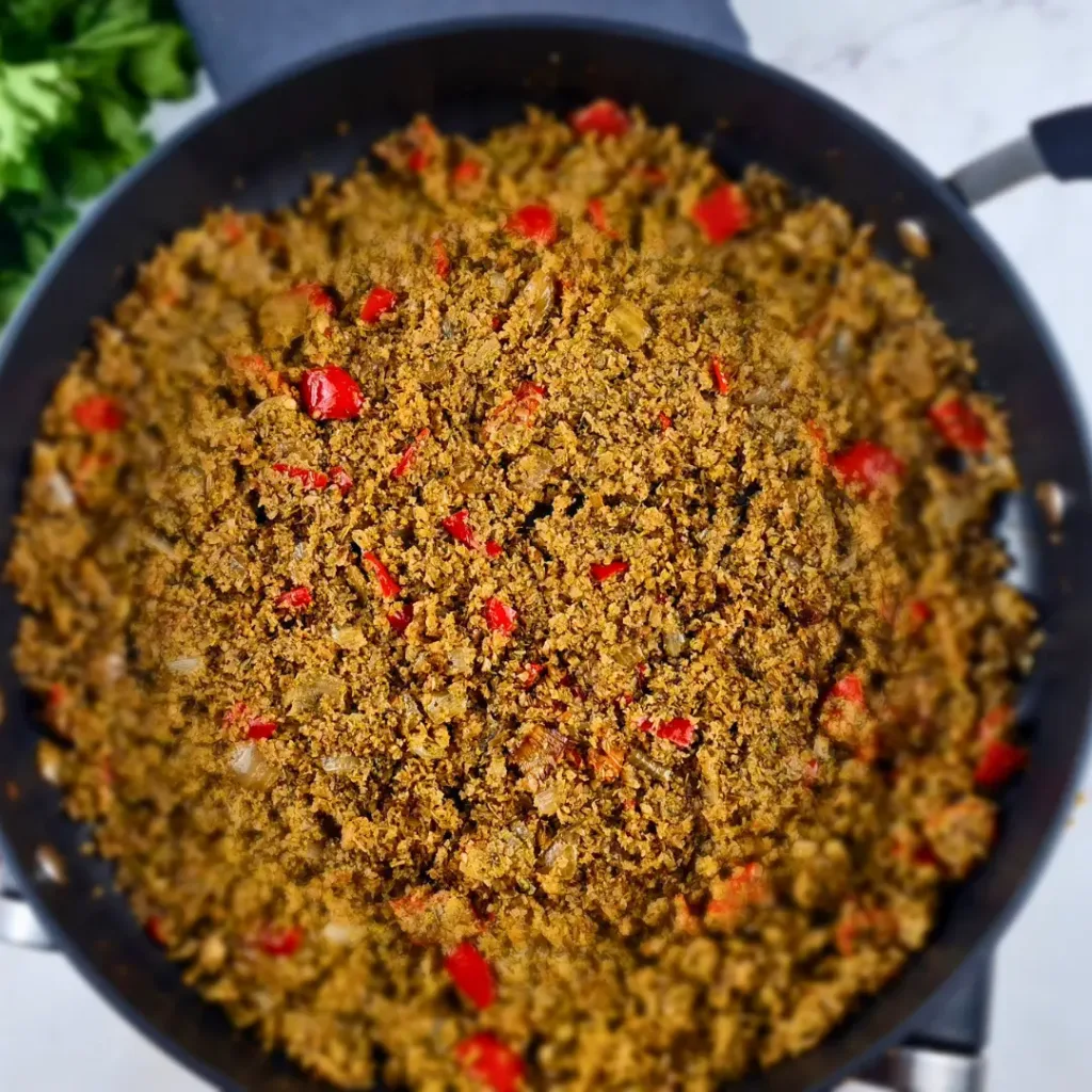 A photo of a black pan filled with a colorful and appetizing dish of Keto Mediterranean Cauliflower & Broccoli Rice, sprinkled with red pepper and onions. The dish is made with cauliflower and broccoli rice, onion, garlic, olive oil, basil, thyme, salt, pepper. It is a low-carb, vegan, and paleo recipe that can be enjoyed as a side dish or a main course. A red pepper is on the left side of the pan, adding some spice and contrast to the dish. The pan is on a white marble countertop, creating a clean and elegant background.