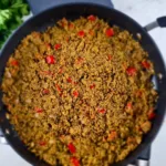 A photo of a black pan filled with a colorful and appetizing dish of Keto Mediterranean Cauliflower & Broccoli Rice, sprinkled with red pepper and onions. The dish is made with cauliflower and broccoli rice, onion, garlic, olive oil, basil, thyme, salt, pepper. It is a low-carb, vegan, and paleo recipe that can be enjoyed as a side dish or a main course. A red pepper is on the left side of the pan, adding some spice and contrast to the dish. The pan is on a white marble countertop, creating a clean and elegant background.