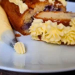 A slice of cake with layers of purple filling and yellow keto royal icing buttercream frosting, next to a piping bag on a white plate.
