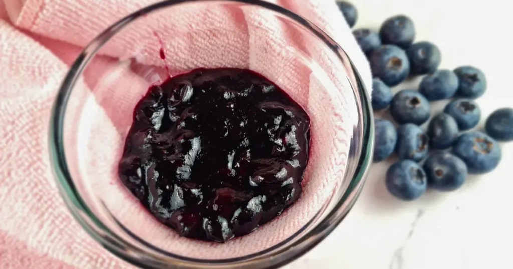 A top view of a bowl of glossy blueberry jam resting on a pink cloth, with fresh blueberries scattered to the side, ready to be used as a topping for keto blueberry cheesecake.