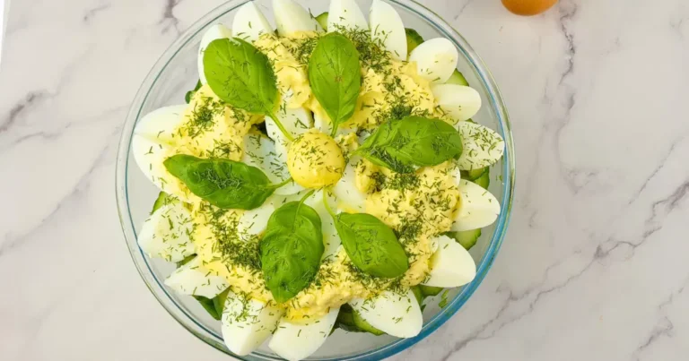 A bowl of keto egg salad with halved boiled eggs, creamy yellow dressing, fresh green basil leaves, and sprinkled herbs on a marbled surface with a whole egg beside it.