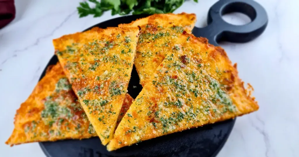 Four slices of golden brown Keto Garlic Bread garnished with herbs, served on a black round cutting board.