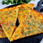 Four slices of golden brown Keto Garlic Bread garnished with herbs, served on a black round cutting board.