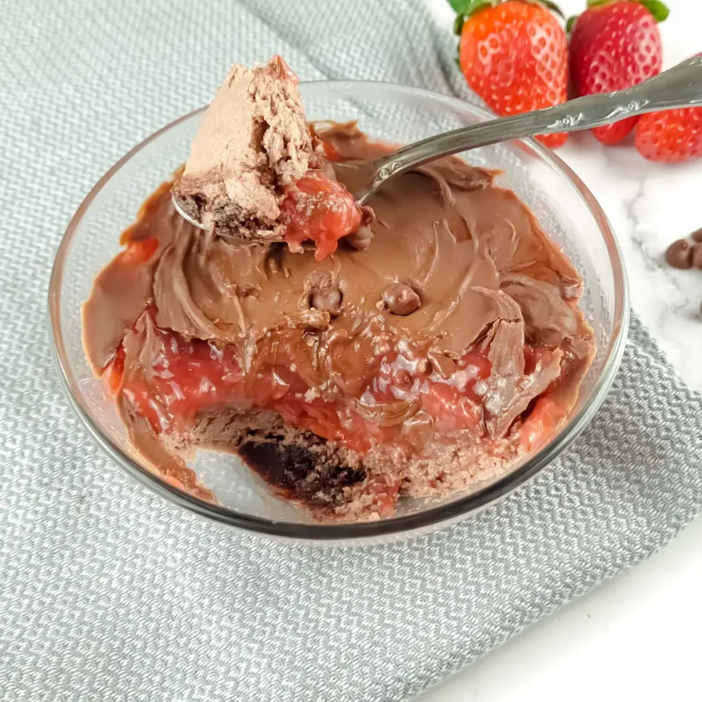 A Keto Mug Cheesecake with chocolate and strawberry jam, fresh strawberries, and chocolate chips on a marble surface.