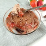 A Keto Mug Cheesecake with chocolate and strawberry jam, fresh strawberries, and chocolate chips on a marble surface.