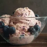 A bowl of high-protein blueberry cottage cheese ice cream, garnished with fresh blueberries on top, presented on a rustic wooden surface with a dark textured backdrop, highlighting the creamy texture and vibrant colors of the dessert.