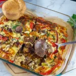 A close-up photo of a baked low carb casserole dish with a golden and creamy topping, filled with chicken liver and vegetables. The keto casserole is garnished with parsley and served on a white plate with a fork. The background is a wooden table with a napkin and a glass of water.