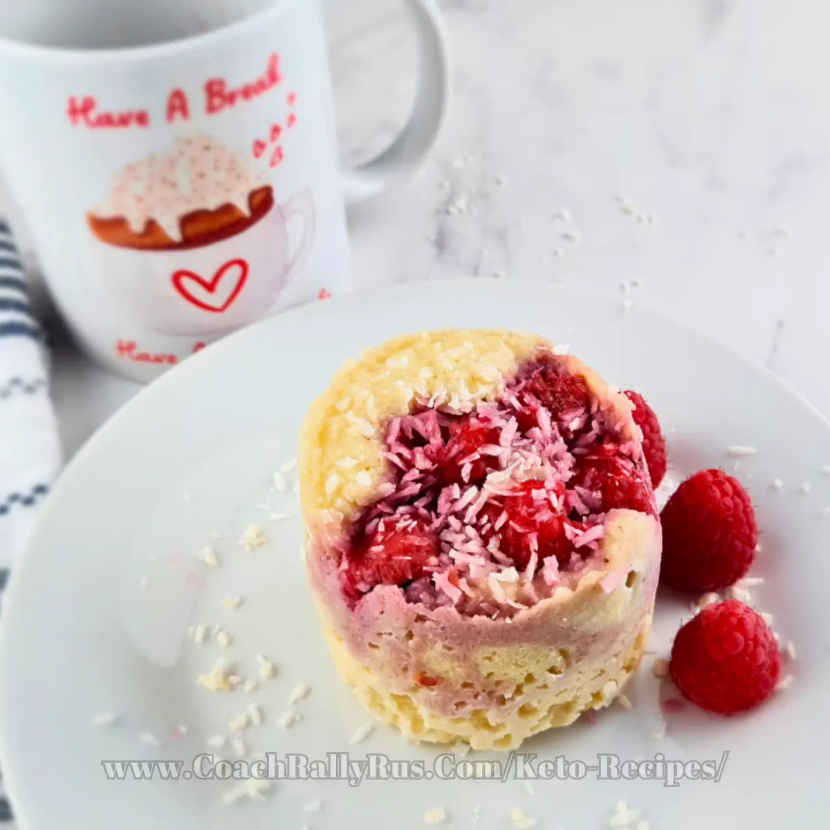 A delicious keto mug cake made with coconut flour and topped with raspberries and shredded coconut, presented on a white plate beside a mug with “Have A Break” text and a donut image, showcasing a tasty yet healthy dessert option.