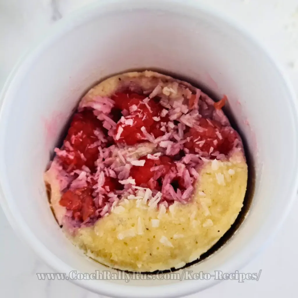A delicious keto mug cake made with coconut flour and topped with raspberries and shredded coconut, presented on a white plate beside a mug with “Have A Break” text and a donut image, showcasing a tasty yet healthy dessert option.