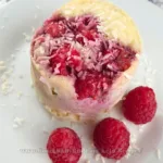A delicious keto mug cake made with coconut flour and topped with raspberries and shredded coconut, presented on a white plate beside a mug with “Have A Break” text and a donut image, showcasing a tasty yet healthy dessert option.