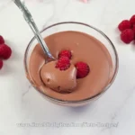Velvety whipped cottage cheese chocolate mousse topped with a fresh raspberry, accompanied by more raspberries on a sleek marble countertop, showcasing a keto-friendly dessert.