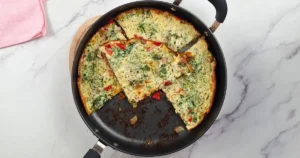 Golden-brown Keto Cottage Cheese Frittata with Pancetta and Broccoli in a black skillet, served on a white marble countertop beside a pink cloth.