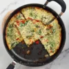 Golden-brown Keto Cottage Cheese Frittata with Pancetta and Broccoli in a black skillet, served on a white marble countertop beside a pink cloth.