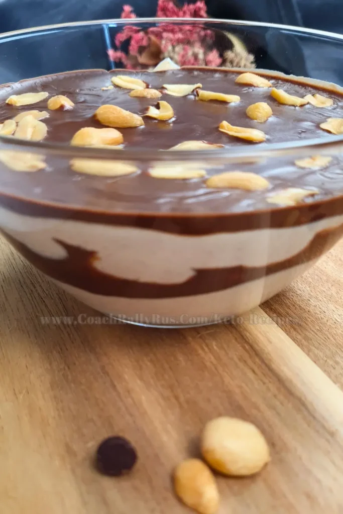 An inviting glass bowl showcases a layered Keto Snickers Mousse dessert with a creamy cottage cheese and peanut butter base, topped with a smooth chocolate sauce and garnished with crunchy whole peanuts, all set against a rustic wooden backdrop with soft-focus dried flowers in the distance.