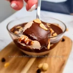 Indulgent Keto Snickers Mousse with layers of smooth cottage cheese and peanut butter blend, topped with a rich chocolate sauce and garnished with whole peanuts, served in a clear glass bowl with a spoonful being lifted, highlighting the dessert’s creamy consistency