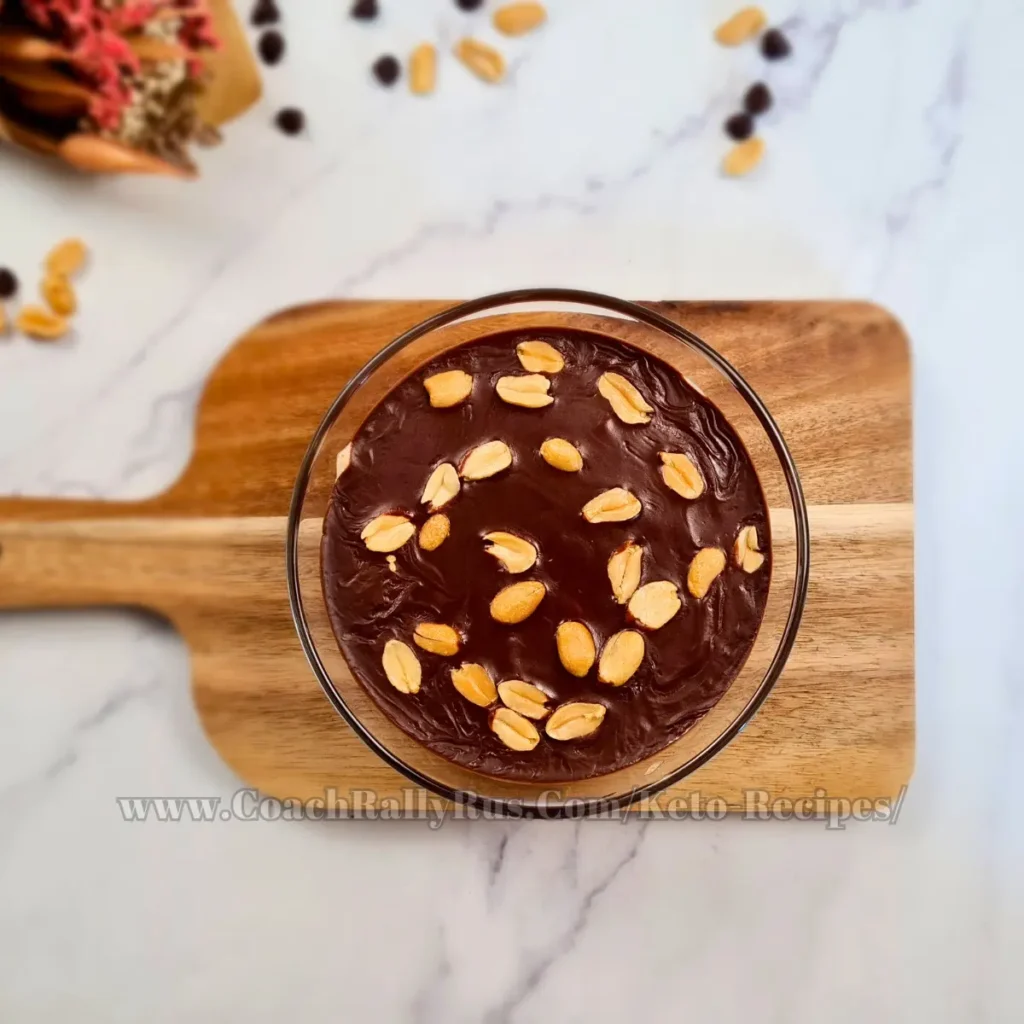 Indulgent Keto Snickers Mousse with layers of smooth cottage cheese and peanut butter blend, topped with a rich chocolate sauce and garnished with whole peanuts, served in a clear glass bowl with a spoonful being lifted, highlighting the dessert’s creamy consistency