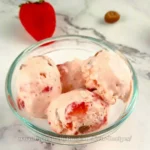 A sumptuous scoop of keto strawberry cheesecake ice cream, garnished with fresh strawberry pieces and crumbled cheesecake crust, served in a stylish glass bowl on a chic marble countertop, embodying a perfect blend of health-conscious indulgence and gourmet dessert presentation.