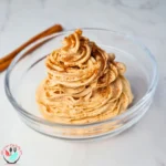 A creamy and light orange-colored pumpkin mousse fluff served in a clear glass bowl, garnished with a sprinkle of dark spices, likely cinnamon or pumpkin spice blend. Two cinnamon sticks lie beside the bowl on a light marbled surface, suggesting the dessert’s flavor profile. This keto, sugar-free, and low-carb dessert is visually appealing and fits dietary restrictions.