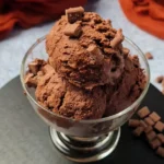 A clear glass bowl filled with scoops of rich Keto Chocolate Ice Cream, presented on a slate coaster. The ice cream has a creamy texture and is garnished with small chocolate chunks. In the background, there’s a soft-focus view of a grey countertop and an orange cloth, which adds a warm contrast to the cool tones of the dessert.