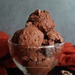 A close-up image of a glass bowl filled with rich, creamy keto chocolate ice cream, garnished with chocolate chips on top. The ice cream has a smooth texture and is set against a dark background with a red cloth partially visible, emphasizing the dessert’s indulgent and keto-friendly qualities.