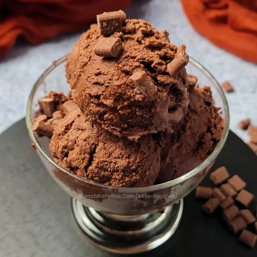 Ninja Creami Ice Cream A clear glass bowl filled with scoops of Ninja Creami Keto Chocolate Ice Cream, garnished with chocolate chunks on top. The bowl is placed on a dark grey surface with a rust-colored cloth in the background. In the corner, there’s a watermark that reads ‘coachallyrus.com/keto-recipes’.