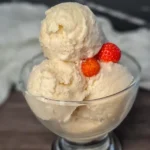 A clear glass bowl filled with two scoops of creamy Ninja Creami Keto Vanilla Ice Cream, garnished with two fresh strawberries on top. The bowl is placed on a dark surface with a white cloth in the background, highlighting the rich texture and pale color of the ice cream, making it an indulgent dessert perfect for a ketogenic diet.