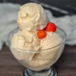 A clear glass bowl filled with two scoops of creamy Ninja Creami Keto Vanilla Ice Cream, garnished with two fresh strawberries on top. The bowl is placed on a dark surface with a white cloth in the background, highlighting the rich texture and pale color of the ice cream, making it an indulgent dessert perfect for a ketogenic diet.