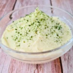 A clear glass bowl filled with creamy Keto Cauliflower Mash, garnished with green herbs, possibly parsley or chives, on a wooden surface. The mash has a smooth texture and is presented as a healthy low-carb alternative to traditional mashed potatoes.