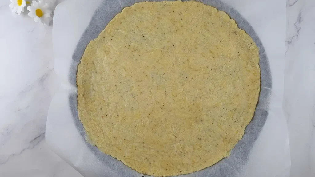 A round almond flour pizza crust is displayed on parchment paper, with a marble countertop in the background. The crust has a golden-brown color and a slightly textured surface, indicating its keto and gluten-free attributes, made from fathead dough. The image is relevant for those interested in low-carbohydrate, gluten-free baking options.