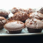 A close-up image of freshly baked coconut flour chocolate muffins with a light dusting of cocoa powder on top, arranged on a white plate against a bright background.