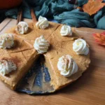 A Cottage Cheese Pumpkin Cheesecake on a wooden surface, with one slice removed, topped with whipped cream and garnished with cinnamon sticks.
