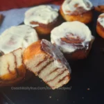A close-up image of freshly baked keto cinnamon rolls made with coconut flour, sitting on a wooden surface. The cinnamon rolls have a golden-brown exterior with visible swirls of cinnamon. Some of the rolls are topped with a white, creamy frosting, adding to their appetizing appearance.