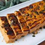 A close-up image of sliced Keto Cottage Cheese Pumpkin Bread topped with pumpkin seeds, presented on a white plate. The bread has a golden-brown crust and a soft, orange-hued interior indicative of pumpkin.